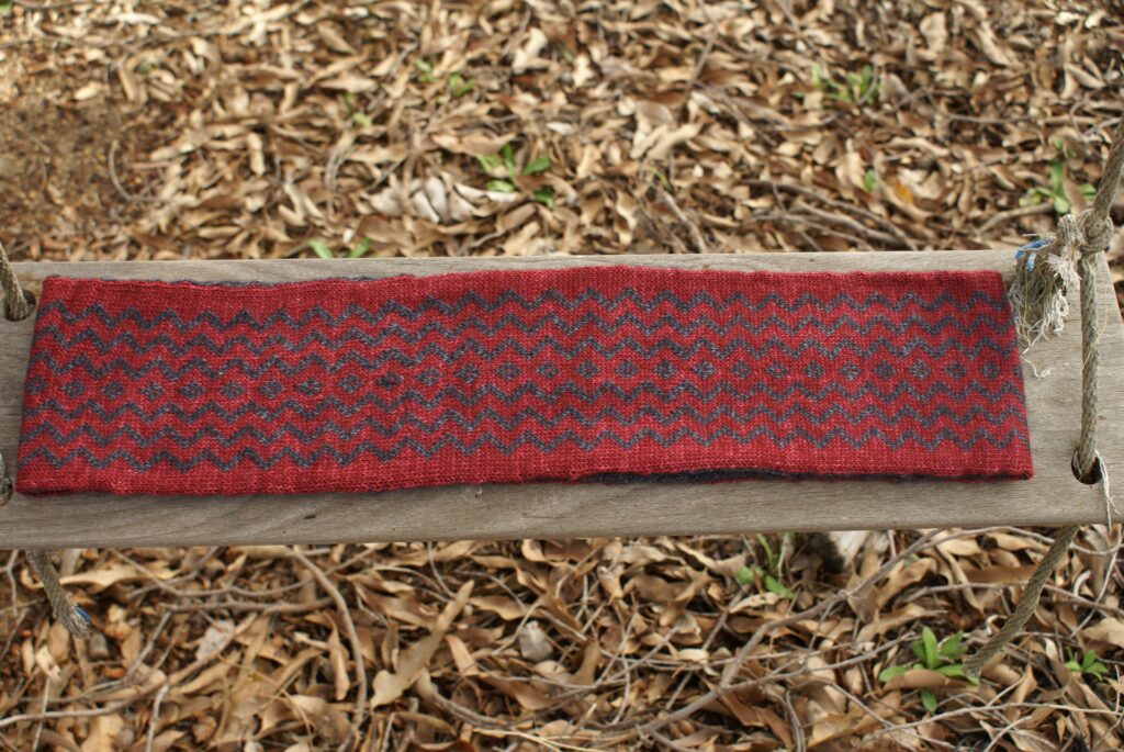 The other side of the cowl on a wooden swing. The cowl has a red background with horizontal charcoal zigzag stripes and a central column of charcoal diamonds.